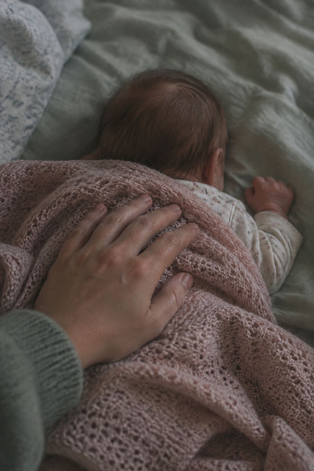A hand resting on a newborn's back beneath a thin knitted blanket in low early-morning light